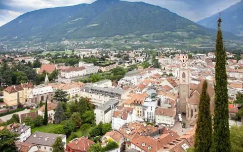 Vista aerea del centro storico di Merano, con montagne sullo sfondo e un campanile che svetta tra i tetti rossi.