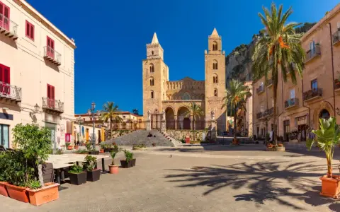 Piazza soleggiata con la cattedrale normanna sullo sfondo, palme e palazzi storici. Un luogo incantevole tra i borghi da visitare in Sicilia.
