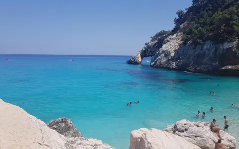 Acqua cristallina e scogliere a picco a Cala Goloritzè, raggiungibile con un suggestivo **percorso a piedi**. Persone nuotano e si rilassano.