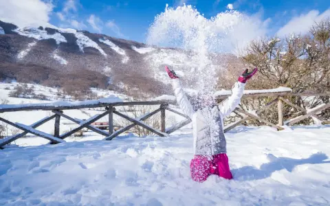 Bambina felice lancia neve in aria, con montagne innevate sullo sfondo. Perfetto per scoprire dove andare per la neve in Abruzzo.