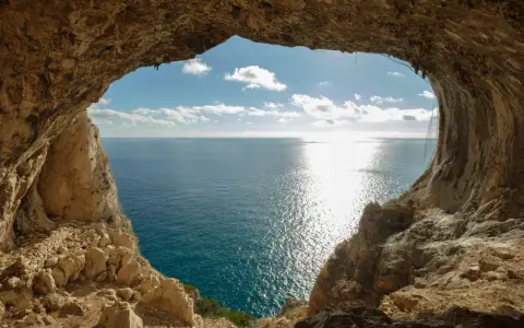 Vista dal mare da una grotta, con un sentiero del pellegrino che si intravede tra le rocce. Il sole brilla sull'acqua.