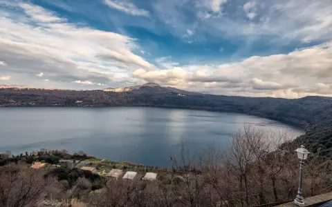 Vista panoramica del lago di Castel Gandolfo, Italia, con colline boscose e un cielo nuvoloso.