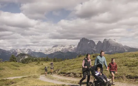 Famiglia in escursione sull'Alpe di Siusi, con bambini e passeggino, sullo sfondo le maestose vette dolomitiche.