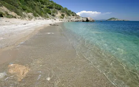 Spiaggia di ciottoli e sabbia con acqua cristallina, vista verso il mare aperto e un'isola in lontananza, vicino al porto di Ercole.