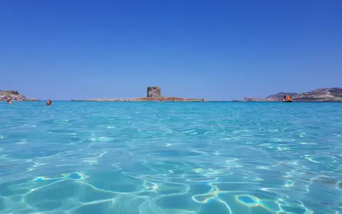 Acque cristalline del mare nord Italia con torre antica su isolotto e barca a vela arancione.