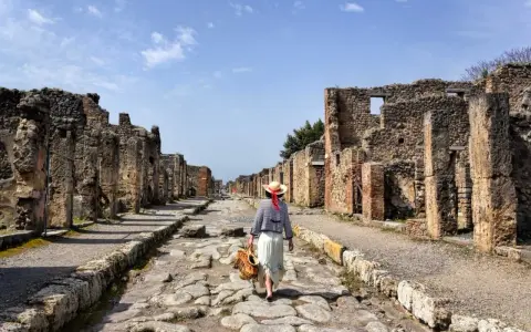 Una donna con cappello di paglia cammina lungo una strada lastricata di Pompei, tra antiche rovine. Un'esperienza unica per scoprire cosa vedere a Pompei.