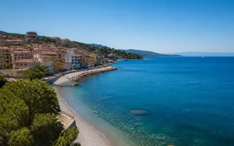 Porto Santo Stefano: case colorate affacciate su spiagge di ciottoli e sabbia, con il mare azzurro e un veliero all'orizzonte.