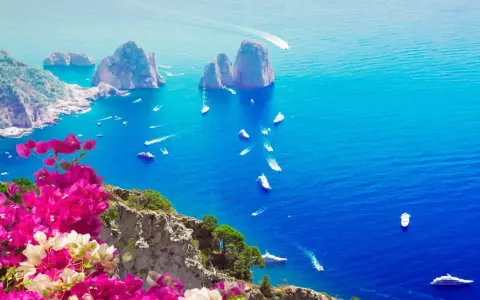 Un giorno a Capri: scogliere rocciose, mare blu intenso e barche che solcano le onde, con bouganville rosa in primo piano.