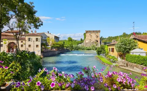 Un fiume attraversa un pittoresco borgo medievale veneto, con un'antica torre in rovina e case colorate.