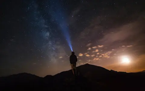 Una persona con una torcia illumina il cielo stellato su una montagna di notte. La Via Lattea è visibile.