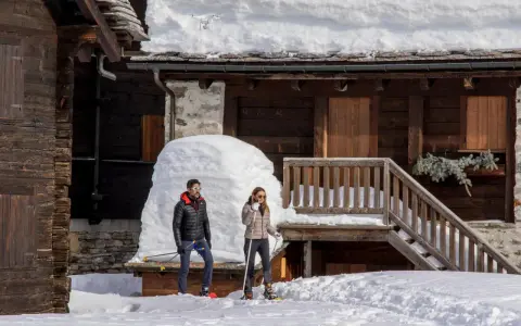 Coppia con racchette da neve a Riale Val Formazza, con un drone in volo sopra un tetto innevato.