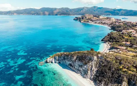 Costa rocciosa con spiaggia bianca e mare cristallino, un borgo fortificato domina la baia. Splendida località mare Toscana.