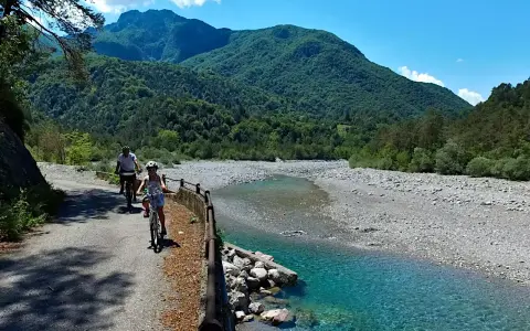 Ciclovia del Tagliamento: due ciclisti esplorano il sentiero panoramico lungo il fiume, circondati da montagne verdi e un cielo azzurro.