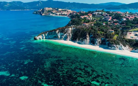 Vista aerea di una spiaggia bianca con acque cristalline e un borgo storico su una collina, parte dell'arcipelago toscano isole visitabili.