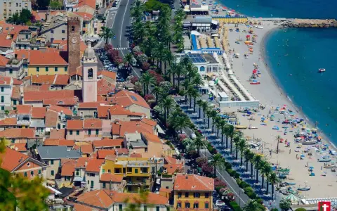 Aerial view of a coastal town with red-tiled roofs, a beach with umbrellas, and palm trees lining the road.