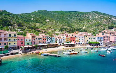 Colorate case affacciate sul mare cristallino, con barche ormeggiate e la collina verde sullo sfondo. Un'immagine che evoca il fascino del traghetto per l'Isola del Giglio.