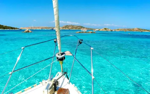 La prua di una barca a vela naviga verso La Maddalena, tra acque cristalline e isole rocciose.
