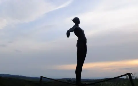 Statua silhouette con braccia aperte, su un crinale collinare, al tramonto. Un punto panoramico lungo le tappe della Via Romea Sanese.