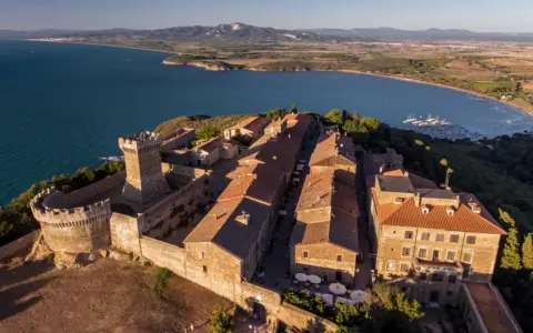 Borgo medievale fortificato domina la campagna toscana lungo il mare, con un castello imponente e case dai tetti rossi affacciate su un golfo con barche a vela.