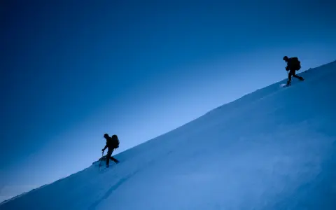 Due escursionisti con ciaspole salgono un pendio innevato sotto un cielo blu intenso. Le ciaspole sono perfette per camminare sulla neve fresca.