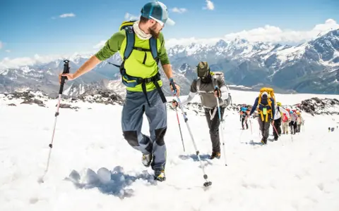 Un gruppo di escursionisti sta camminando sulla neve in montagna, con bastoncini da trekking e zaini, sotto un cielo azzurro.
