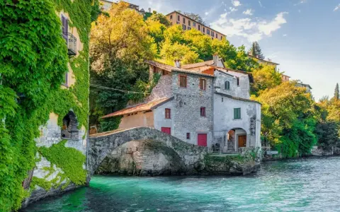 Antichi borghi medievali lombardia da visitare: un ponte di pietra collega case storiche sull'acqua cristallina, con edera rampicante e vegetazione lussureggiante.
