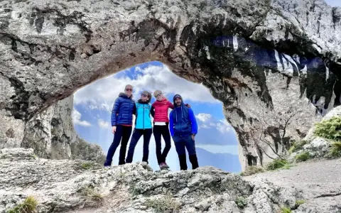 Quattro amici si godono una giornata di camminare in montagna, posando sotto un arco naturale di roccia con un cielo azzurro e nuvoloso.