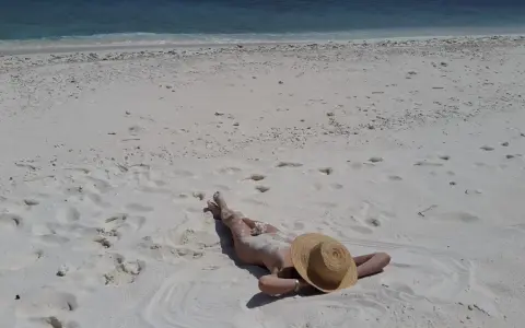 Persona distesa su sabbia bianca, con cappello di paglia, vicino al mare turchese. Un'immagine che evoca la pace delle spiagge naturiste in Italia.