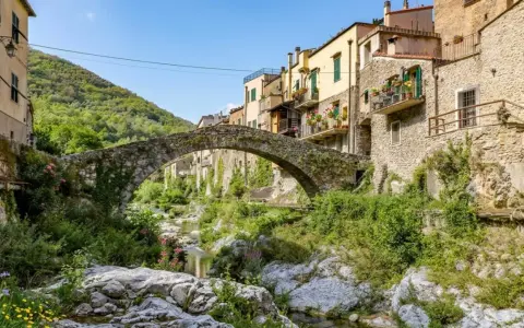 Antico ponte in pietra collega i borghi dell'entroterra ligure ponente, con case colorate e vegetazione rigogliosa che scende verso il torrente.
