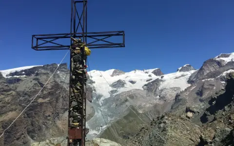 Croce metallica e di pietra in cima a una montagna, con un ghiacciaio e vette innevate sullo sfondo. Il **palon di Resy** domina il panorama.