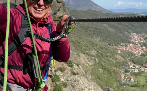 Donna sorridente si aggrappa a un cavo d'acciaio, godendosi un'avventura sulle ferrate Val di Susa con un panorama mozzafiato.