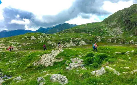 Escursionisti attraversano un paesaggio verdeggiante in una località montagna Piemonte, con rocce e nuvole minacciose all'orizzonte.