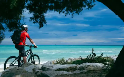 Ciclista ammira il mare turchese lungo la ciclovia adriatica in Abruzzo, con alberi che incorniciano la scena.