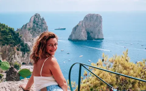 Donna sorridente con vista sui Faraglioni, un'ispirazione per come visitare Capri. Mare azzurro e barche in lontananza.