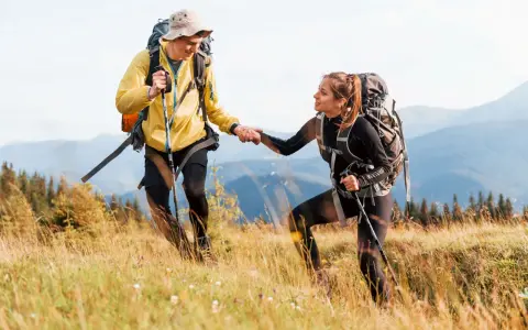 Coppia di escursionisti si aiutano a vicenda su un sentiero di montagna, con zaini e bastoncini da trekking.