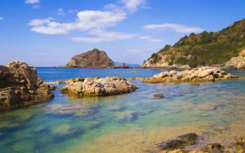 Acque cristalline e rocce scolpite dal mare a Porto Ercole, con montagne verdi sullo sfondo e un cielo azzurro punteggiato di nuvole.