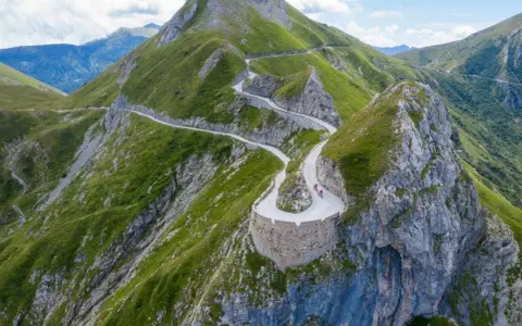 Un tortuoso cammino del sale si snoda tra montagne verdi, con ciclisti che affrontano tornanti mozzafiato su un sentiero panoramico.