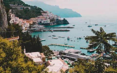 Vista panoramica di un villaggio colorato sulla Costiera Amalfitana, con barche nel porto e montagne sullo sfondo. Un invito a scoprire come visitare la Costiera Amalfitana.