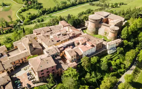 Aerial view of a medieval Italian village with a castle, terracotta roofs, and surrounding green hills.