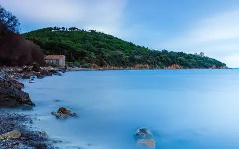 Una casa di pietra sulla spiaggia di Bengodi, con il mare calmo e una collina verdeggiante sullo sfondo.