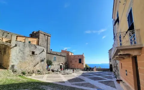 Piazza nel centro storico di Borgio Verezzi con edifici antichi, tavolini all'aperto e il mare sullo sfondo.