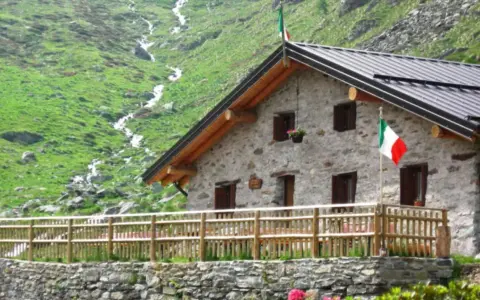 Rifugio in pietra con bandiera italiana, circondato da verdi pendii e un ruscello. Un'oasi accogliente tra i rifugi Val Camonica facili da raggiungere.