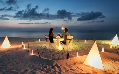 Una coppia al mare di sera, seduta a tavola per una cena romantica illuminata da lanterne e luci soffuse sulla sabbia.