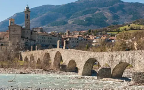Ponte medievale in pietra attraversa un fiume, con un borgo storico e colline sullo sfondo. Un gioiello tra i borghi medievali dell'Emilia Romagna.