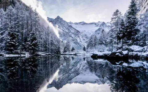 Val di Mello inverno: alberi innevati e montagne maestose si specchiano in un lago calmo, creando un paesaggio da fiaba.