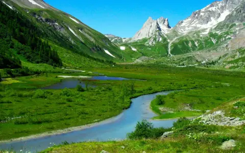 Val Ferret, un paradiso per passeggiate facili. Un fiume serpeggia tra prati verdi e montagne innevate sotto un cielo azzurro.