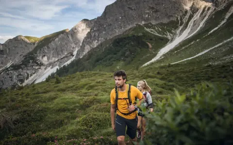 Coppia in escursioni alto adige, camminano su un sentiero verdeggiante con montagne imponenti sullo sfondo.