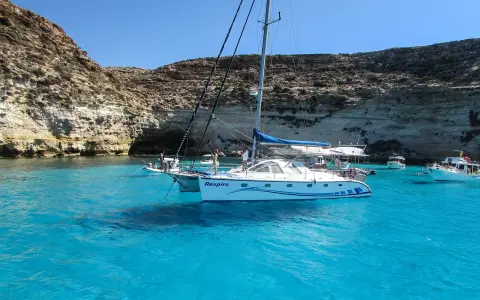 Catamarano "Respiro" in acque cristalline di un'isola sotto la Sicilia, con scogliere rocciose e altre barche.