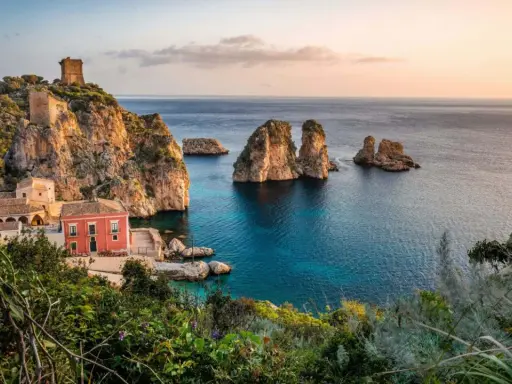 Un borgo con torri antiche e una casa rossa si affaccia su un mare bello Italia, con faraglioni che emergono dalle acque cristalline.