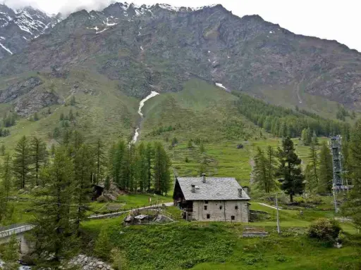 Un casolare in pietra nel **Val di Rhemes**, circondato da verdi pendii e imponenti montagne innevate.